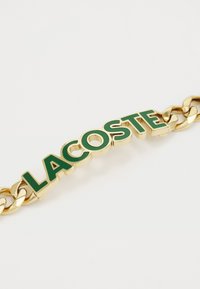 Bracelet en chaîne dorée avec un logo "LACOSTE" en émail vert, possédant une texture lisse et des maillons épais pour une durabilité accrue.