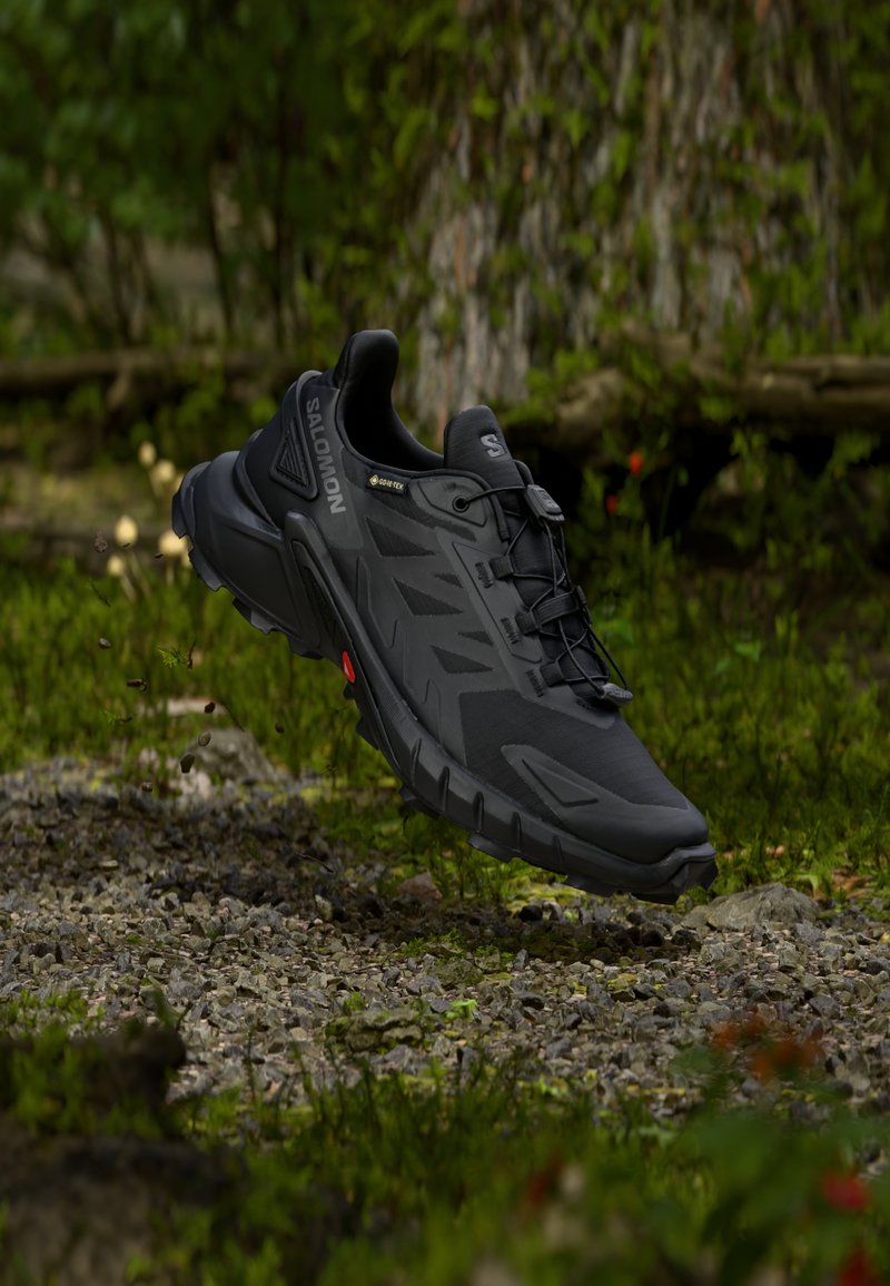 Čierna trailová bežecká obuv Salomon Gore-Tex vznášajúca sa vo vzduchu nad skalnatou lesnou pôdou s rozmazaným zeleným listnatým pozadím.