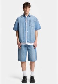 Svijetloplava traper košulja s kratkim rukavima i prednjim džepovima, u kombinaciji s odgovarajućim traper šortsevima i crnim tenisicama. Bijele čarape su vidljive.