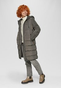 Doudoune grise avec capuche, présentant une fermeture éclair et des sections matelassées horizontales. Portée par-dessus un pull crème et un jean gris, avec des bottines beige.