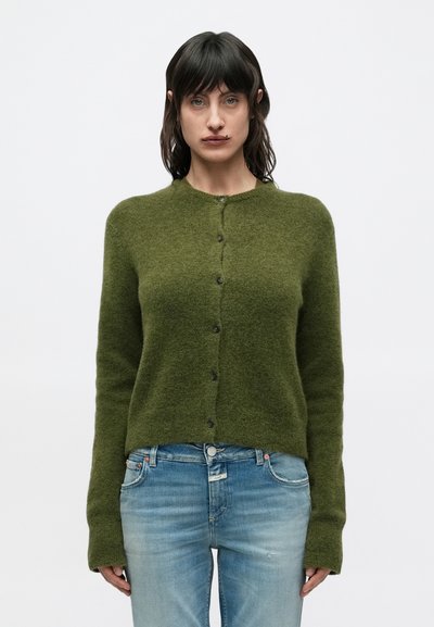 Femme portant un cardigan en maille vert olive boutonné et un jean bleu clair, debout devant un fond blanc uni.
