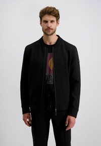 Schwarze Zip-Jacke mit rundem Halsauschnitt, aus glattem Stoff. Mit gerippten Bündchen und einem Logo-Patch auf dem linken Ärmel. Getragen über einem bedruckten T-Shirt.