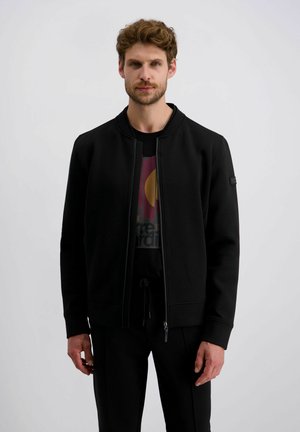 Zwarte vest met rits en ronde halslijn, gemaakt van gladde stof. Voorzien van ribgebreide manchetten en een logo patch op de linker mouw. Gedragen over een graphic tee.