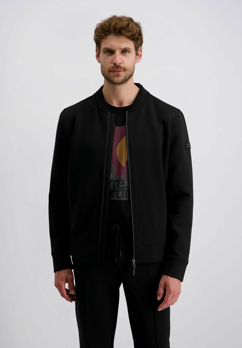 Schwarze Zip-Jacke mit rundem Halsauschnitt, aus glattem Stoff. Mit gerippten Bündchen und einem Logo-Patch auf dem linken Ärmel. Getragen über einem bedruckten T-Shirt.
