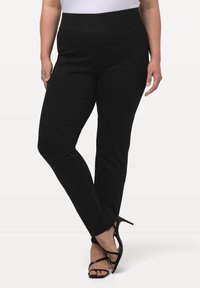 Donna che indossa pantaloni neri attillati e sandali con tacco alto neri, in piedi con le gambe incrociate su uno sfondo neutro.