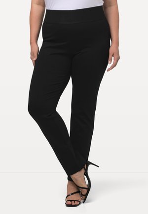 Donna che indossa pantaloni neri attillati e sandali con tacco alto neri, in piedi con le gambe incrociate su uno sfondo neutro.