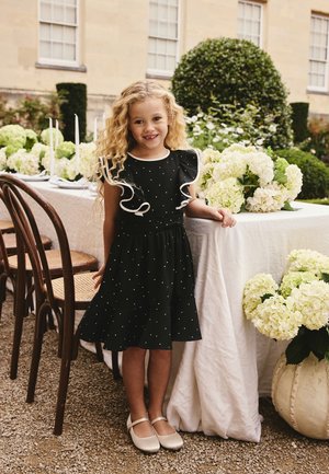 Jong meisje met krullend haar dat een zwarte jurk met witte rand draagt, staat naast een met bloemen versierde tafel buiten bij een beige gebouw.