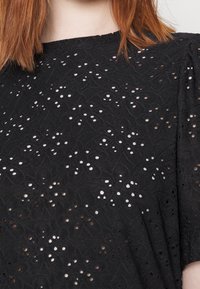 Černý top se složitými výřezy ve tvaru oka, s malými volánovými rukávy a texturovaným povrchem. Kulatý výstřih a volný střih.