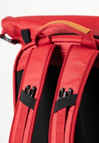 Zaino rosso con spallacci imbottiti e maniglia superiore. Include accessori in metallo nero e dettagli in pelle marrone chiaro a contrasto.