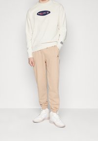 Ljusgräddfärgad sweatshirt med "NIKE"-logga, kombinerad med beige sweatpants med sidrand och vita höga sneakers.