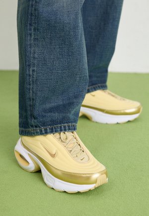 Jambes en jean bleu et baskets Nike jaune pâle à semelle blanche debout sur un sol vert clair.