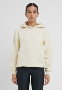 Cremefarbene Kapuzenjacke mit einer Fronttasche und weißen grafischen Text. Weicher Stoff, lockere Passform, mit Kapuze und gerippten Bündchen. Kombiniert mit schwarzen Leggings.