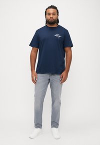 Μπλε ναυτικό βαμβακερό T-shirt με κοντά μανίκια, που έχει ένα μικρό τυπωμένο λογότυπο στο αριστερό στήθος. Συνδυάζεται με ανοιχτό γκρι τζιν και λευκά αθλητικά παπούτσια.