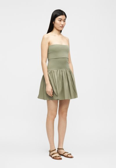 Νεαρή γυναίκα με μακριά σκούρα μαλλιά, ντυμένη με ελιάς πράσινο φόρεμα χωρίς τιράντες και καφέ επίπεδα σανδάλια, στέκεται μπροστά σε απλό φωτεινό φόντο.