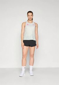 Frau steht vor einfarbigem Hintergrund, trägt ein weißes ärmelloses Sportoberteil, schwarze Shorts, weiße Socken und weiße Turnschuhe.