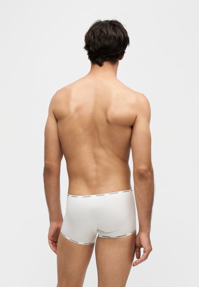 Jeune homme aux cheveux foncés portant un boxer blanc, debout dos à la caméra sur un fond uni.