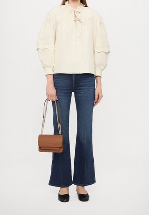 Frau trägt eine cremefarbene Bluse mit Puffärmeln, blaue ausgestellte Jeans, schwarze Ballerinas und hält eine braune Lederhandtasche mit goldenen Kettenträgern.