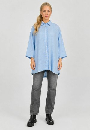 Femme aux cheveux blonds portant une chemise oversize bleu clair boutonnée, un jean gris droit et des bottines noires, debout face à nous.