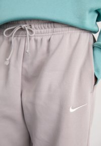 Lys grå sweats med elastisk blomst og snøre, hvid Nike swoosh logo, iført med en teal langærmet top, hånd i lommen.