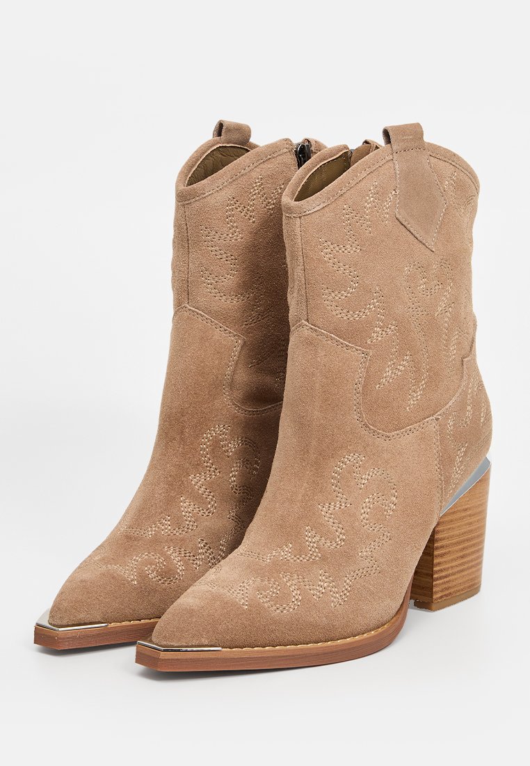 Botas de vaquero de ante marrón claro con puntas afiladas, costuras decorativas y un tacón de madera grueso. Cuenta con un cierre de cremallera en la parte trasera.