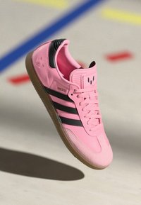 Rózsaszín sportcipő fekete csíkokkal, kerek orral, sima textúrával, fűzős kivitele és gumi talpa van. Az oldalán "MESSI" felirat található.