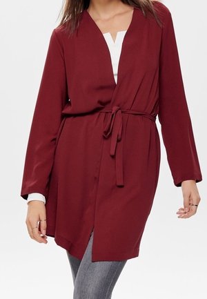 Frau trägt eine weinrote Wickel-Strickjacke mit langen Ärmeln, die an der Taille gebunden ist, über einem weißen Oberteil und grauen Jeans, steht mit leicht gebeugten Armen.