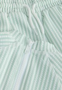 Světle zelená mikina na zip s proužkovaným vzorem a bílými akcenty. Vyrobena z texturované látky, s elastickými manžetami a pasem.