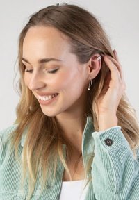 Boucles d'oreilles en argent avec un accent en perle, portées par une femme aux longs cheveux ondulés, vêtue d'une veste en velours côtelé vert clair. Expression souriante.