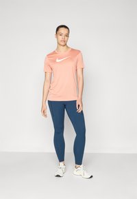 Nike lyhythihainen paita vaalean korallin värissä, jossa valkoinen logo, yhdistettynä sinisiin legginsseihin. Ominaisuuksiin kuuluu sileä pinta ja istuva muotoilu.