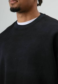 Zwarte sweater met een gestructureerd oppervlak, voorzien van kleine zwarte kralen die gelijkmatig verspreid zijn. Geribde ronde hals met daaronder een gelaagd wit overhemd.