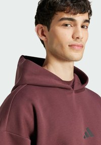 Bordeaux huppari, valmistettu pehmeästä kankaasta, jossa on sileä rakenne. Etuosassa on musta Adidas-logo ja kaksinkertainen huppu.