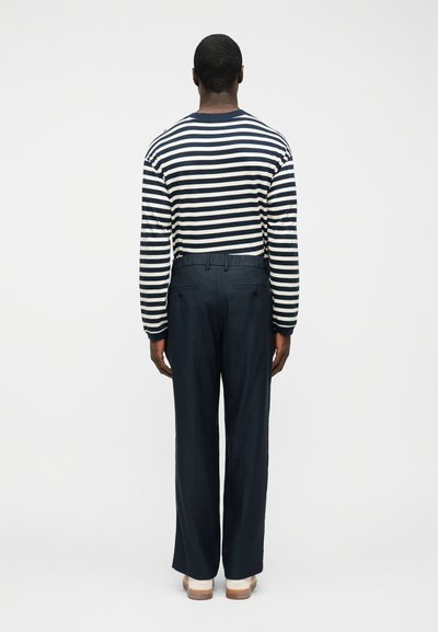 Άνδρας που στέκεται με την πλάτη γυρισμένη φοράει μαύρη και άσπρη ριγέ μακρυμάνικη μπλούζα, σκούρο μπλε παντελόνι και λευκά παπούτσια σε απλό λευκό φόντο.