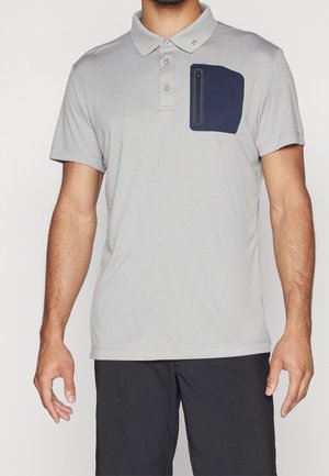 Hombre con una camiseta tipo polo gris claro de manga corta y un bolsillo en el pecho con cremallera azul oscuro, combinado con pantalones negros.
