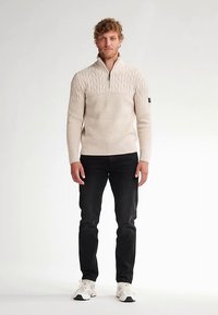 Hellbeiger, gerippter Pullover mit Reißverschlusskragen und strukturierter Musterung im oberen Bereich. Kombiniert mit schwarzen Jeans und weißen Sneakers.