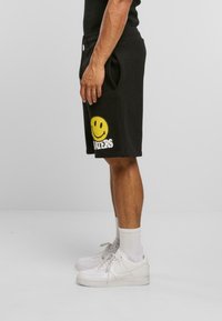 Sorte sweatshorts med sidelommer, med et stort gult smiley-ansigt grafisk design og hvid tekst "HADERS" på venstre ben.