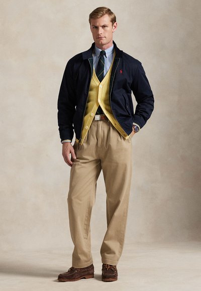 Bărbat purtând o jachetă bleumarin, cardigan galben, cămașă albastră cu cravată în dungi, pantaloni bej, pantofi maro și ceas de mână, stând în fața unui fundal simplu.