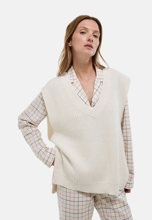 Gilet en tricot beige clair avec un décolleté en V, tissu texturé et épaules tombantes, associé à une chemise à carreaux et un pantalon à motifs.
