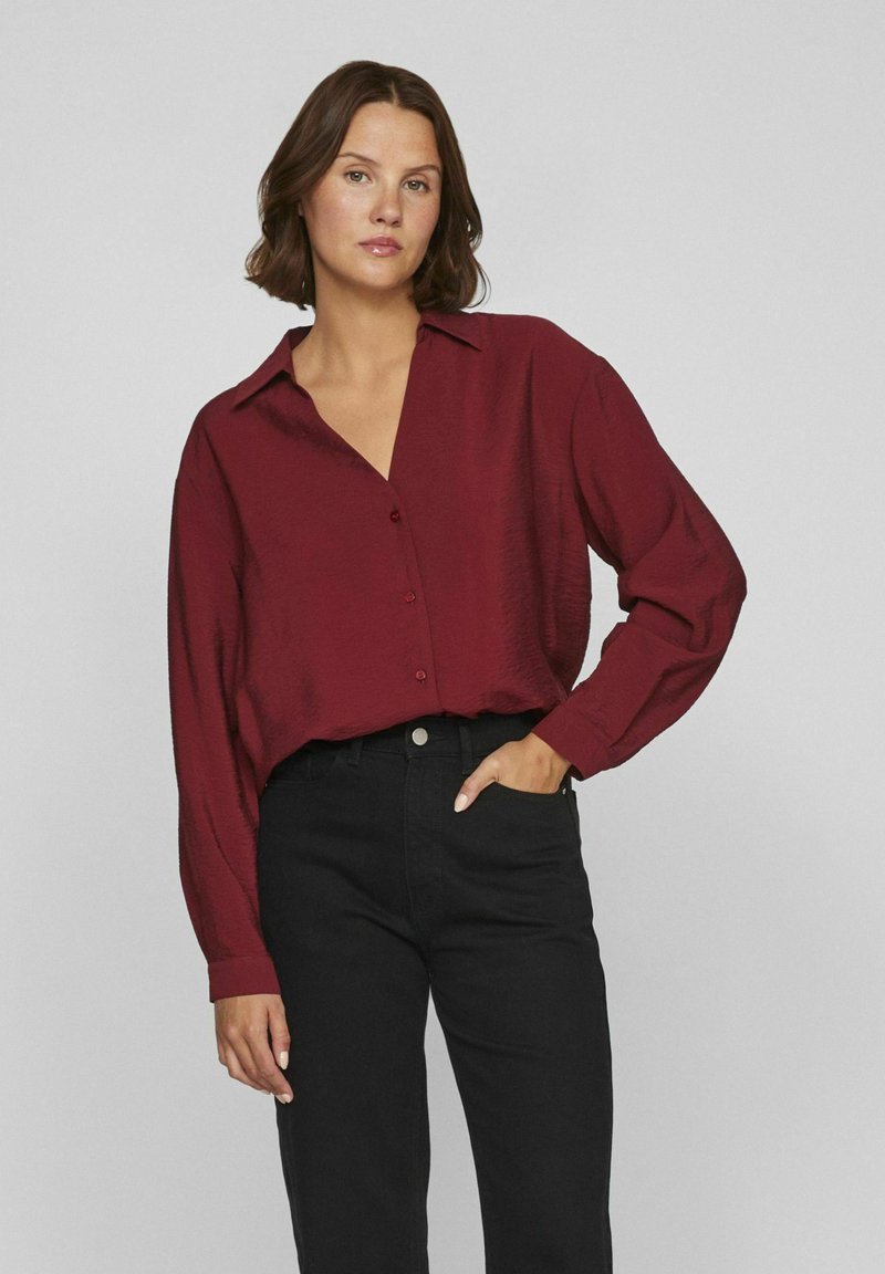 Camisa burdeos de manga larga con cuello, cierre de botones y ajuste relajado, combinada con jeans negros de tiro alto. Acabado de tela texturizada.