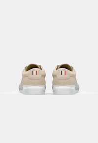 Beige semskinn-sneakers med hvit gummisåle, med en glatt finish og en liten dekorativ rød, hvit og blå stripe på hælen.