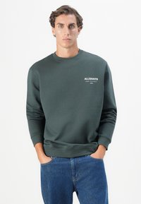 Sweatshirt verde com punhos e bainha canelados, apresentando um pequeno logótipo branco no peito, combinado com jeans azuis. Textura suave, design casual.