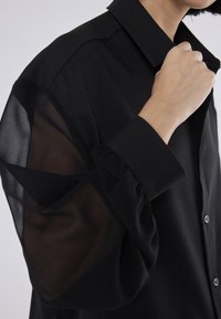 Personne ajustant le col d'une chemise noire aux manches longues transparentes et aux poignets opaques sur un fond uni.