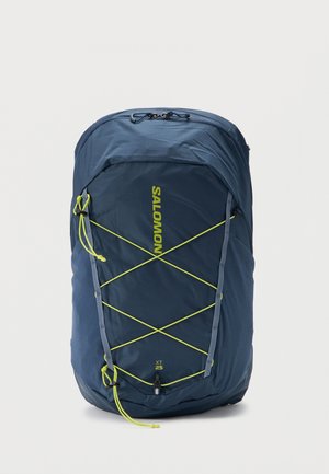 Sac à dos Salomon XT 25 bleu foncé avec des cordons bungee jaunes en forme de croix et des poches latérales en mesh sur fond blanc.