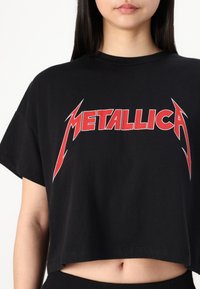 Černé zkrácené tričko vyrobené z bavlny, s výrazným červeným textem "METALLICA" v nápadném písmu. Krátké rukávy a volný střih.