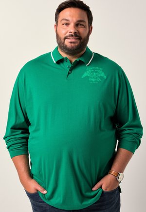 Grün langärmliges Polohemd mit weißem Kragen und bedrucktem Logo, kombiniert mit dunklen Jeans. Weicher Stoff, entspannte Passform, lässiges Design.