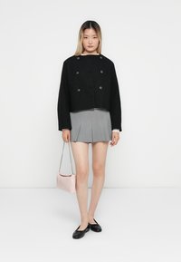 Veste noire courte avec double boutons, jupe plissée grise, ballerines noires et un sac à main rose avec une bandoulière en chaîne. Fond épuré.