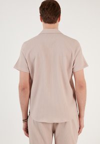 Camisa de manga corta de color rosa claro, con textura acanalada y un corte relajado. La camisa presenta un collar clásico y un dobladillo redondeado.