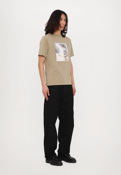 Camiseta de algodón beige con un estampado gráfico cuadrado en colores suaves, combinada con pantalones negros con patrón y calzado negro.