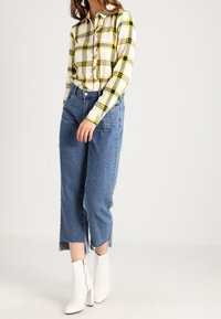 Camisa de cuadros amarilla y blanca con botones, combinada con unos jeans azules de cintura alta. La modelo lleva botas de tobillo blancas con tacón y un acabado texturizado.