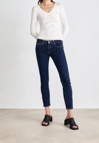 Top blanco de manga larga con escote en V, combinado con jeans ajustados de color azul oscuro. Los jeans presentan una textura de mezclilla y detalles de herrajes estándar. Mule de tacón cuadrado en color negro.