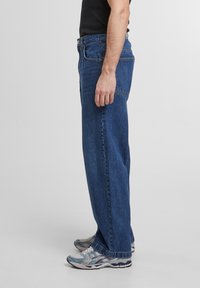 Jeans en denim bleu foncé, coupe décontractée avec poches latérales. Portés avec des chaussures de sport grises et bleues. T-shirt noir uni visible au-dessus de la taille.